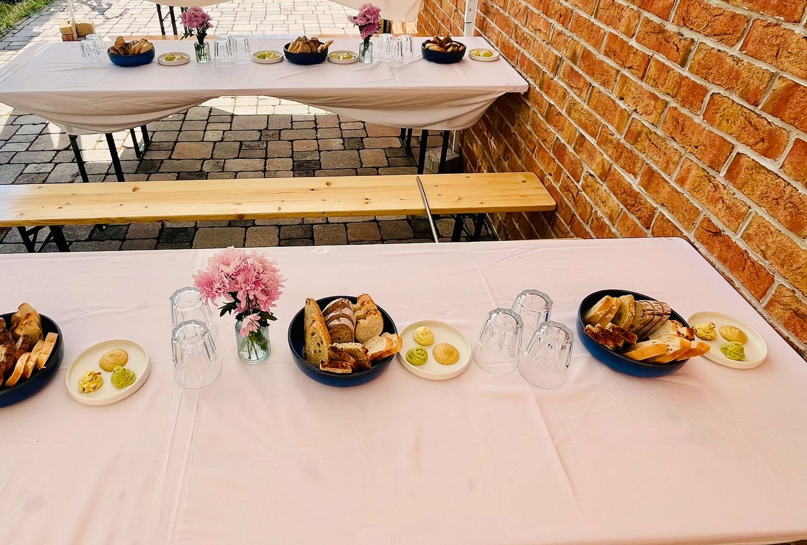Im Freien aufgebaute lange Tafeln mit Brotkörben, rosa Blumen und Gläsern vor einer Backsteinwand