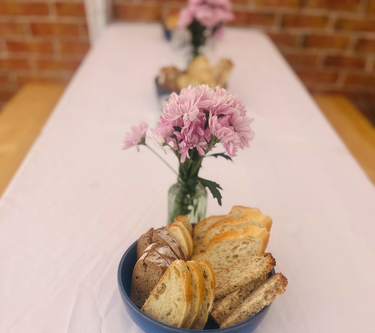 Lange weiß gedeckte Tafel vor einer Backsteinwand, darauf Brotkörbe und rosa Blumen in kleinen Vasen