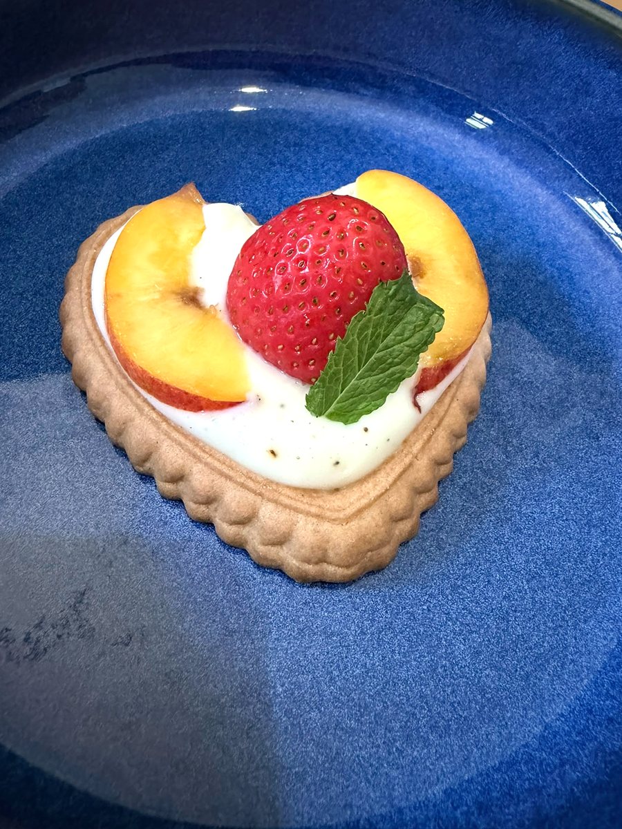 Herzförmiges Mürbeteig-Törtchen mit Creme, Erdbeere, Pfirsich und Minze auf tiefblauem Teller